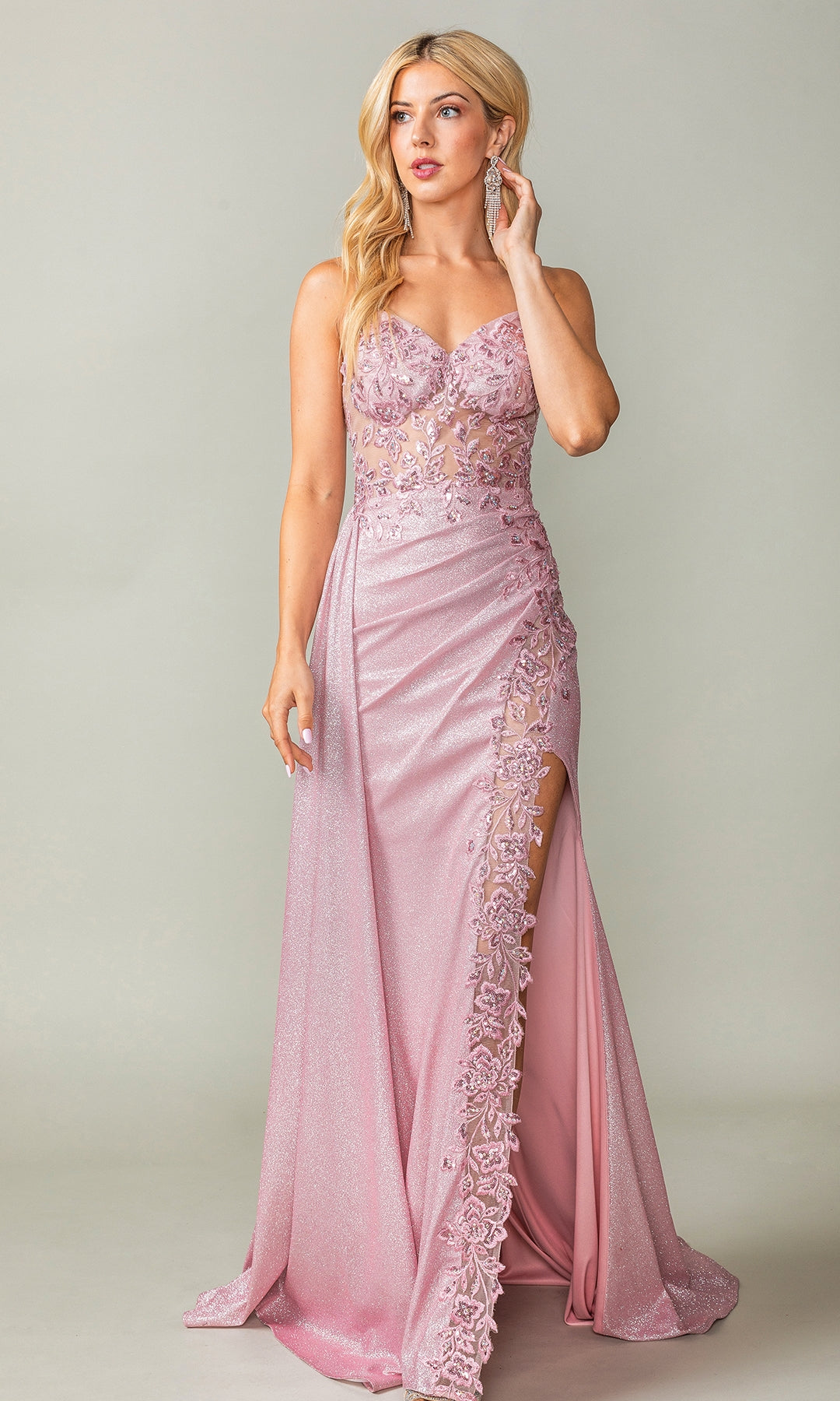A woman models the Draped Long Glitter Prom Dress: Dancing Queen 4377, featuring a shimmery pink hue, corset-style bodice, lace details, thigh-high slit, and spaghetti straps against a plain light background.